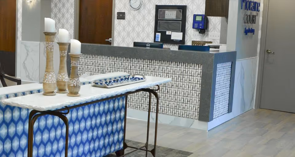 Reception area of Moraine Court Supportive Living featuring a front desk with a patterned front panel, a console table with three decorative candle holders and candles, a wall-mounted clock, and a keypad device. The floor has light wood flooring and the walls have a geometric patterned wallpaper.