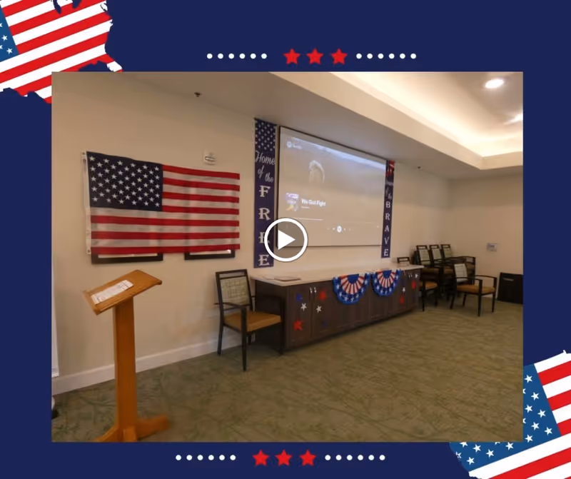 Interior room decorated with patriotic American flags and banners. A large American flag is mounted on the wall next to a wooden podium and a chair. A cabinet decorated with red, white, and blue bunting and stars is beneath a projector screen displaying a video titled 'We Got Fight'. Several stacked chairs are visible in the corner of the room.