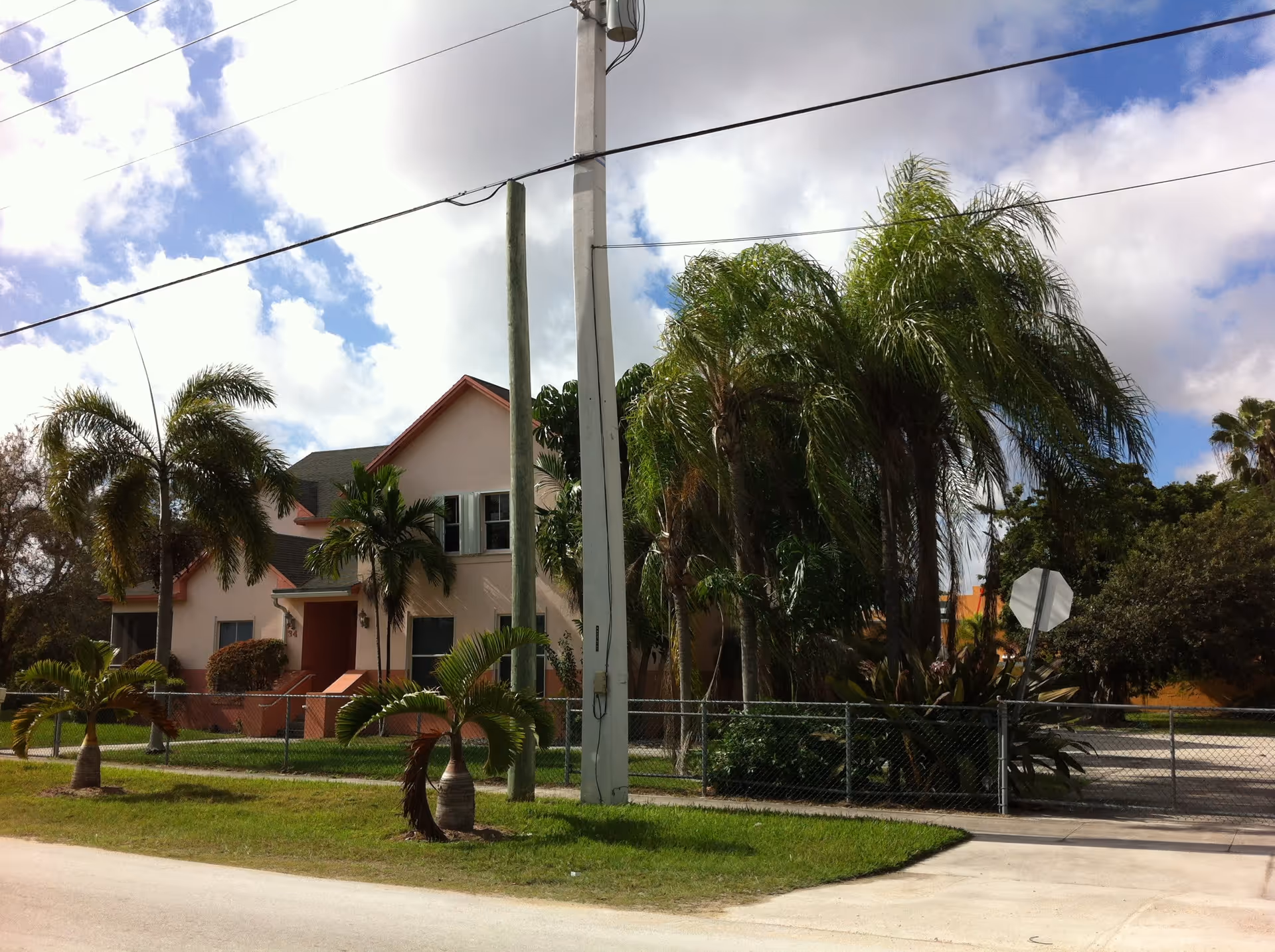 Exterior view of a two-story residential building with a light pink facade and red trim, surrounded by palm trees and greenery. There is a chain-link fence enclosing the property and a utility pole with power lines in front. The sky is partly cloudy with patches of blue.