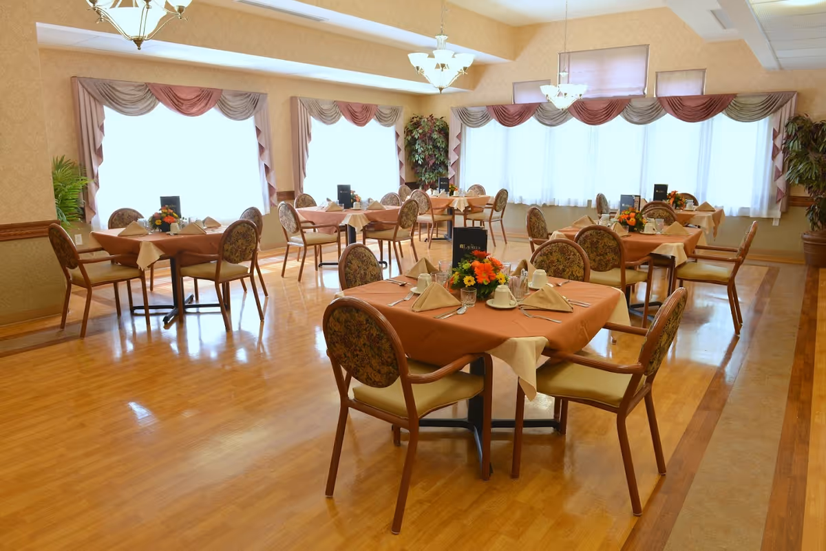 Bright dining room with multiple round tables set with orange tablecloths, floral centerpieces and chairs near large windows.