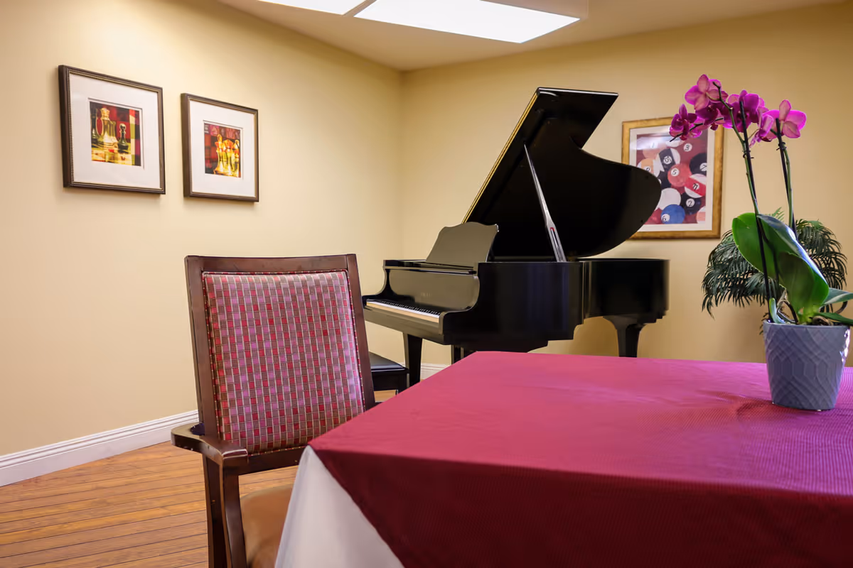 Interior common room with a black grand piano, a table covered by a red tablecloth and an orchid, and a chair against a beige wall with framed artwork.