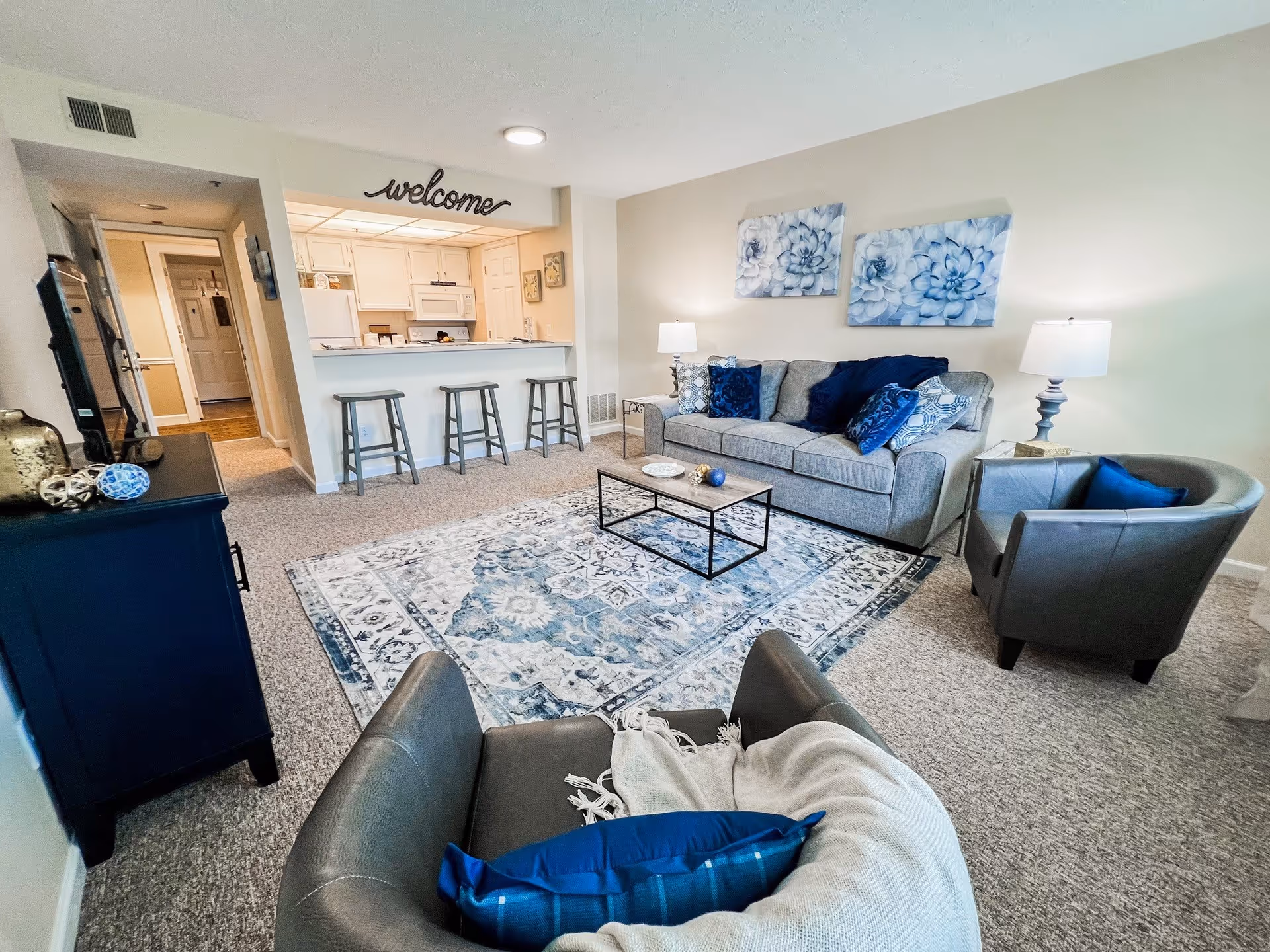 A bright and cozy living room with a gray sofa adorned with blue and patterned cushions, two gray armchairs, a wooden coffee table on a blue and white patterned rug, and two floral wall art pieces above the sofa. The room opens to a kitchen area with white cabinets, a microwave, and a refrigerator. Three gray bar stools are placed at the kitchen counter. A 'welcome' sign is displayed above the kitchen opening.