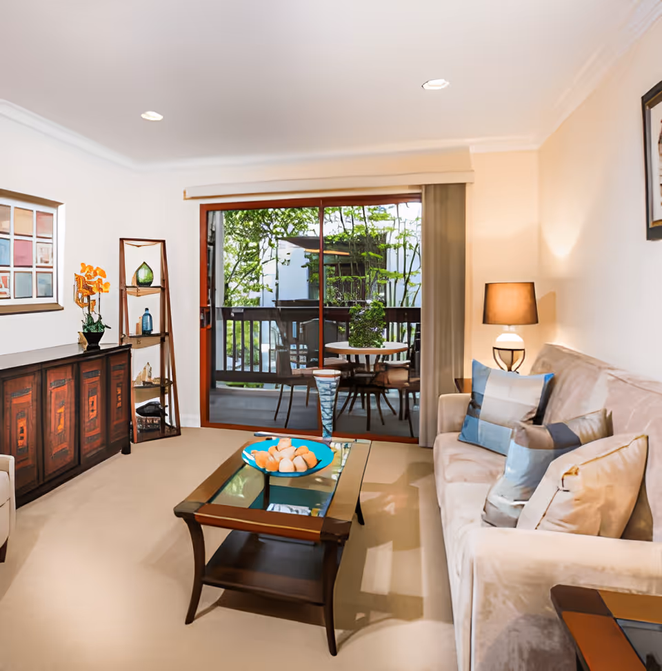 Bright living room with a sofa, coffee table, shelving and sliding glass doors leading to an outdoor patio.