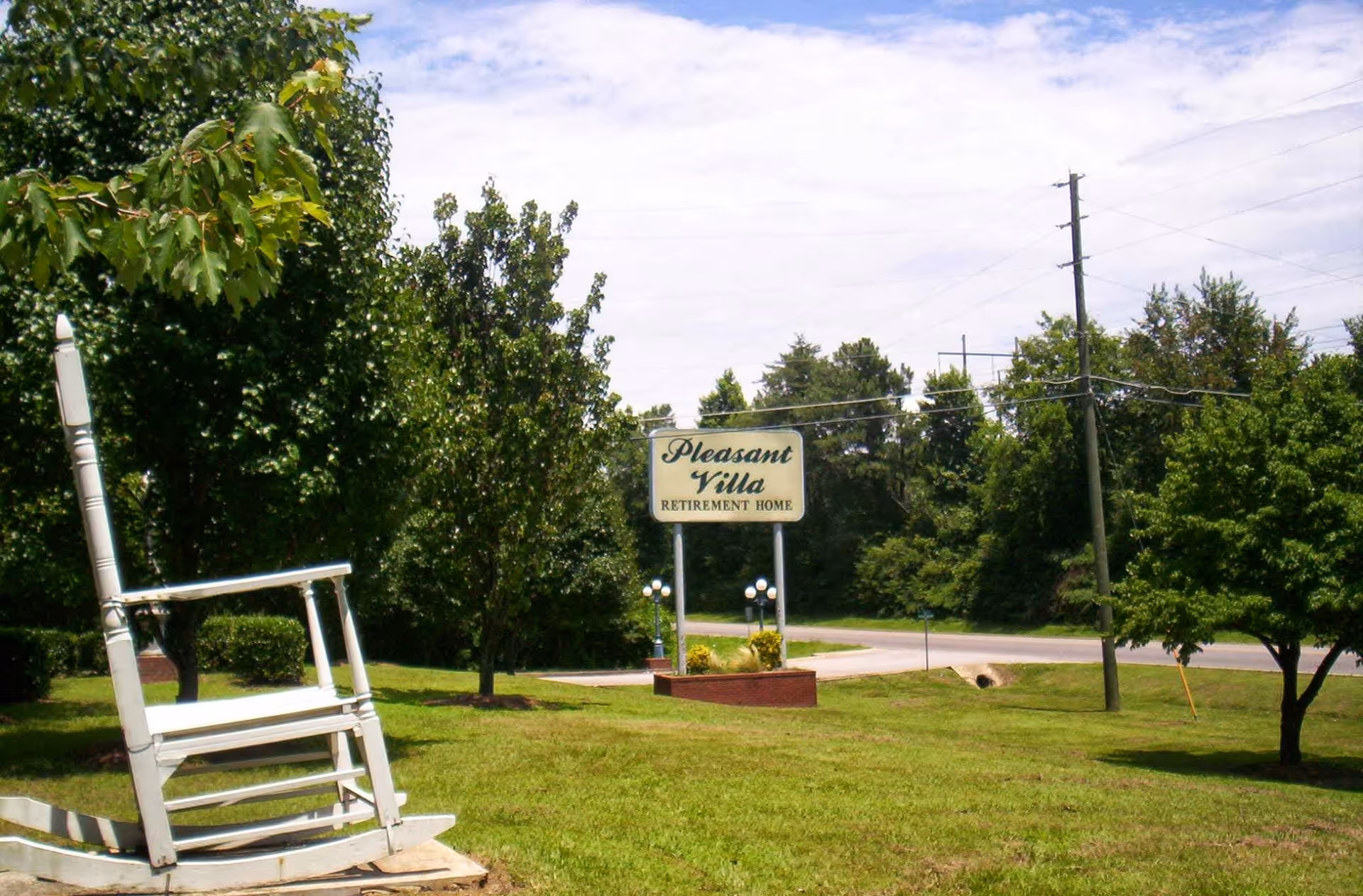 Outdoor view of Pleasant Villa Retirement Home sign on a grassy area with trees and a large white rocking chair sculpture in the foreground. A road and utility poles are visible in the background under a partly cloudy sky.