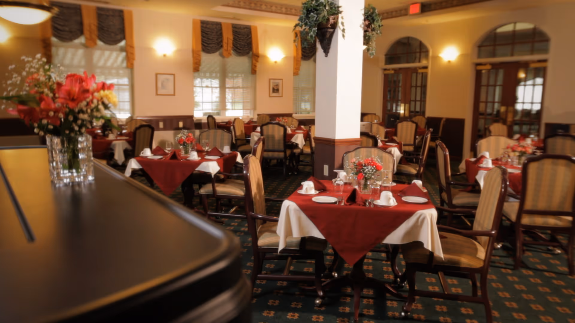 A dining room with multiple tables covered in white tablecloths and red overlays, each set with plates, cups, and napkins. The room features upholstered chairs, floral centerpieces on the tables, warm lighting, and windows with decorative curtains.