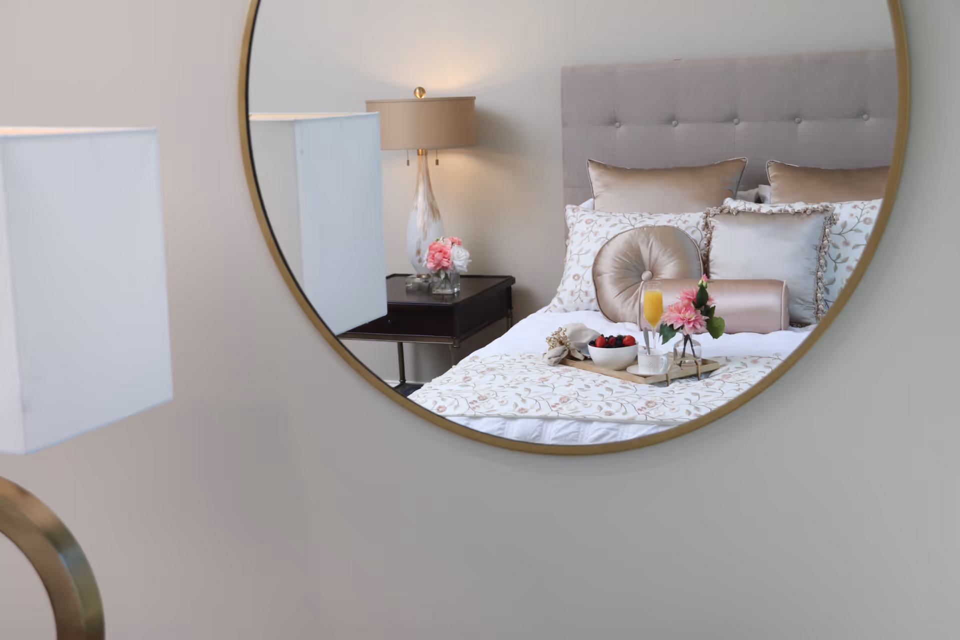 Round mirror reflects a neatly made bedroom with decorative pillows, a bedside table lamp and flowers, and a breakfast tray on the bed.