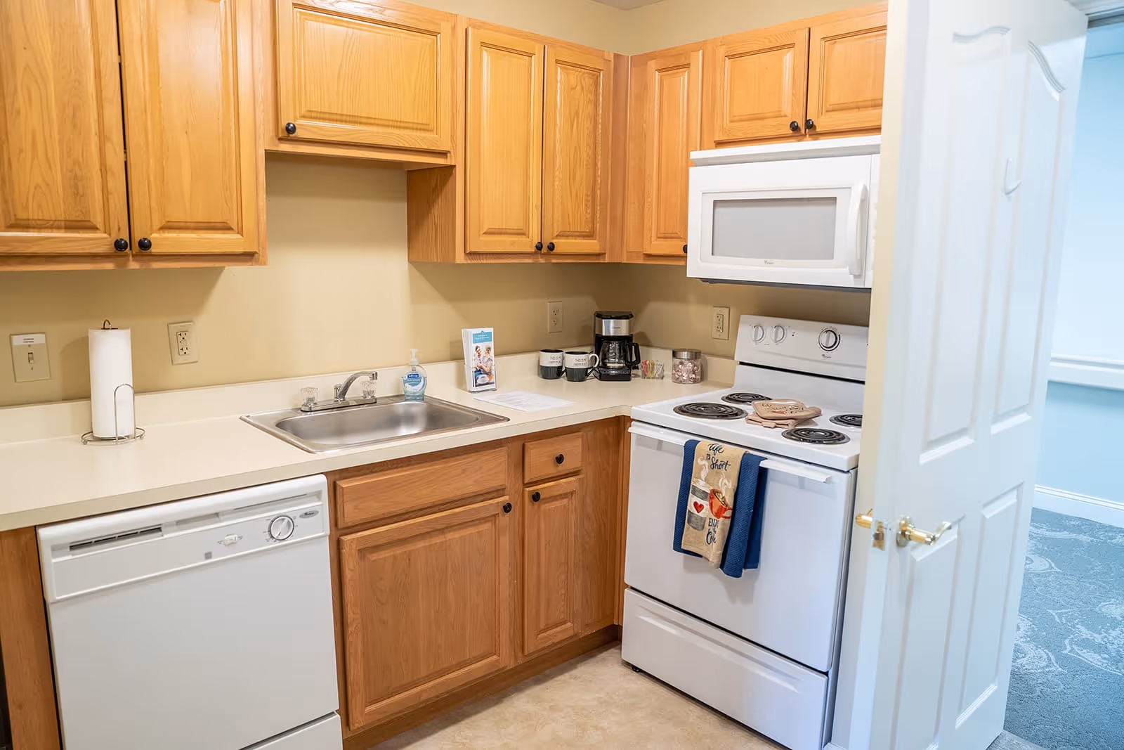 A small kitchen area with wooden cabinets, a white dishwasher, a stainless steel sink, a white electric stove with oven, and a white microwave above the stove. There are kitchen towels hanging on the oven handle, a coffee maker, mugs, and a paper towel holder on the countertop. A partially open white door reveals a carpeted room beyond.