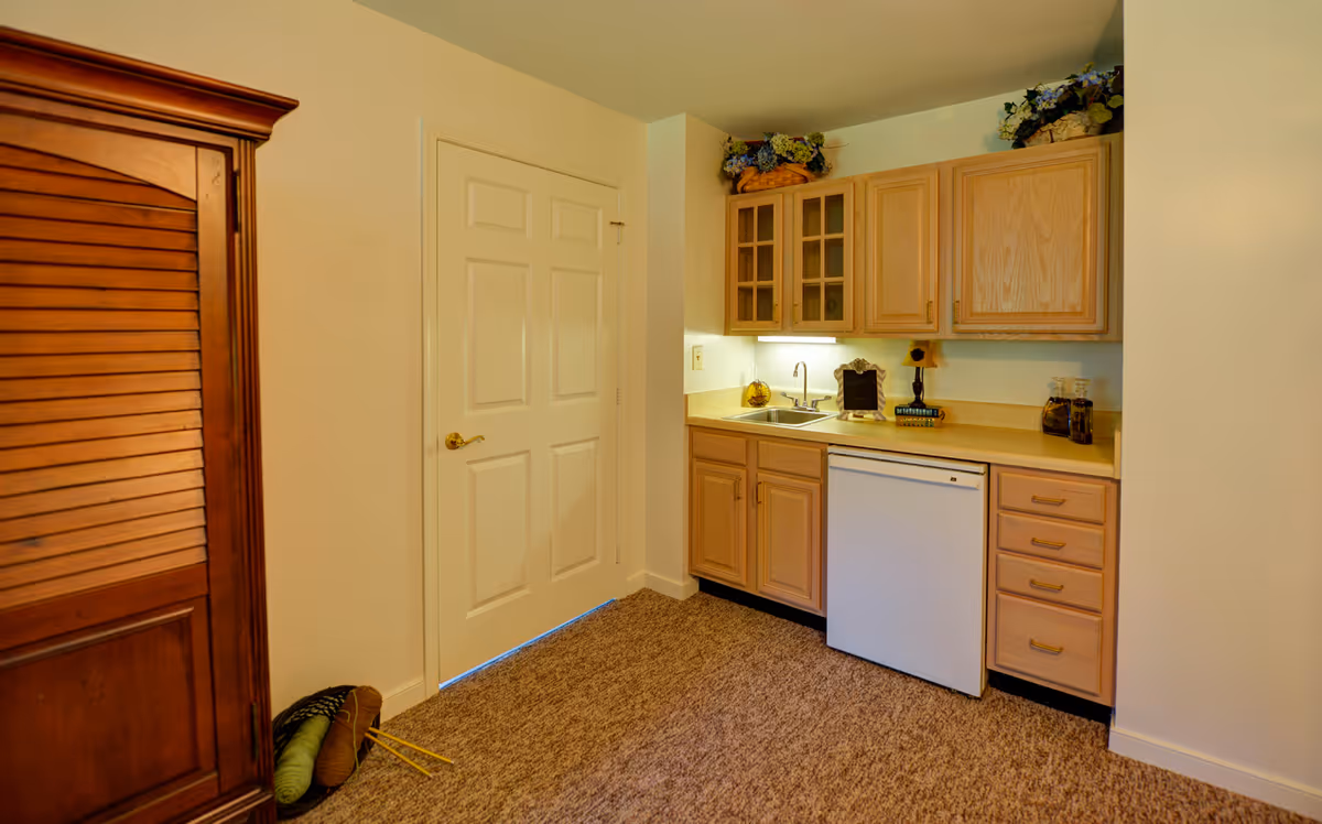 A small kitchenette area with light wood cabinets, a sink, a mini refrigerator, and countertop decorations including a small lamp and a chalkboard. To the left is a closed white door and a wooden armoire. The floor is carpeted and the walls are painted light beige.