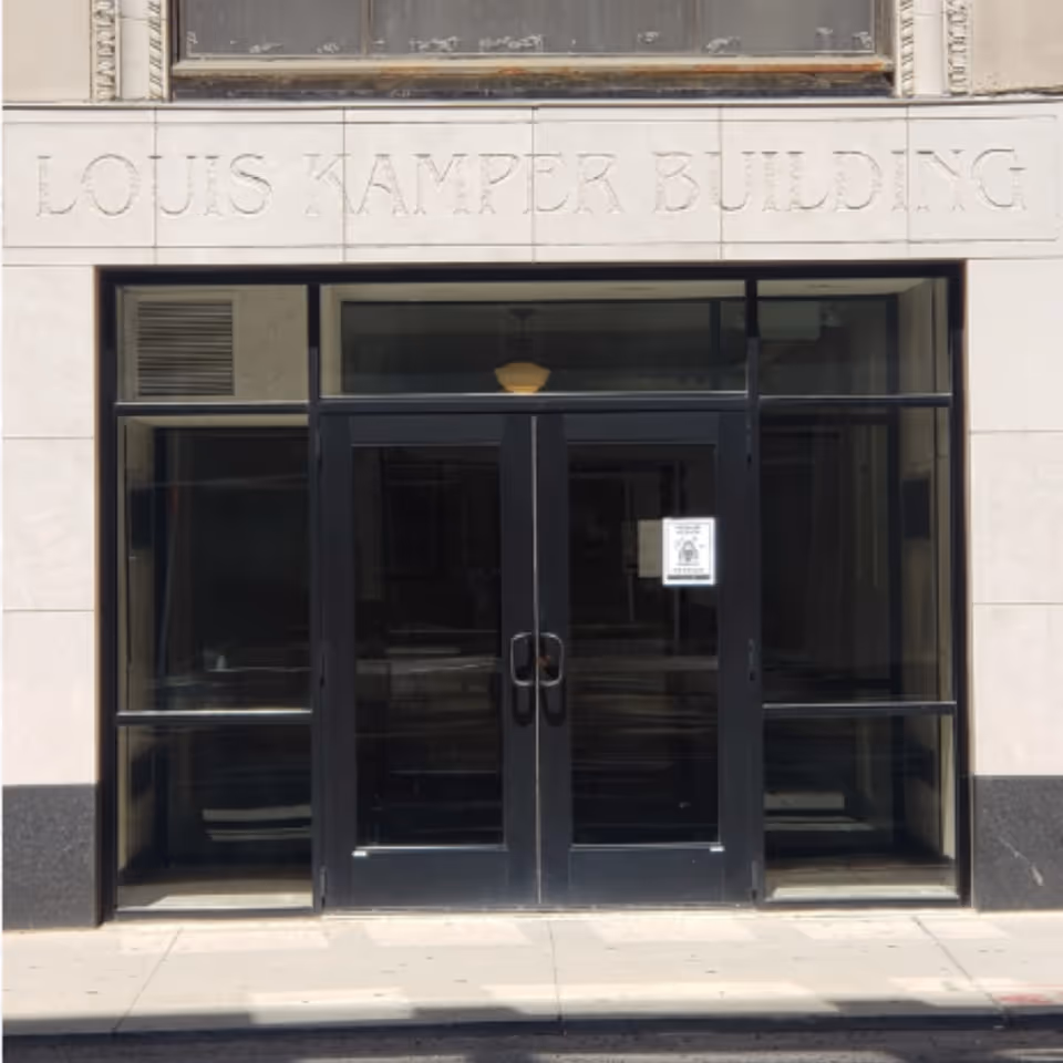 Entrance to the Louis Kamper Building featuring double black framed glass doors with a sign posted on one door, surrounded by light-colored stone walls and a sidewalk in front.