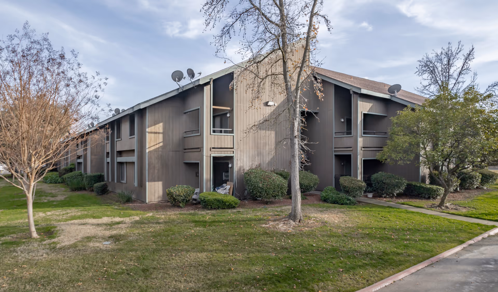 Exterior view of a two-story brown building with balconies and satellite dishes on the roof, surrounded by grass, trees, and bushes under a partly cloudy sky.