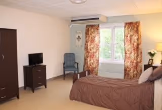 A cozy bedroom in a senior living facility with a bed covered in a brown comforter, a small dark wooden dresser with a TV on top, a blue upholstered chair, and a window with floral curtains letting in natural light.
