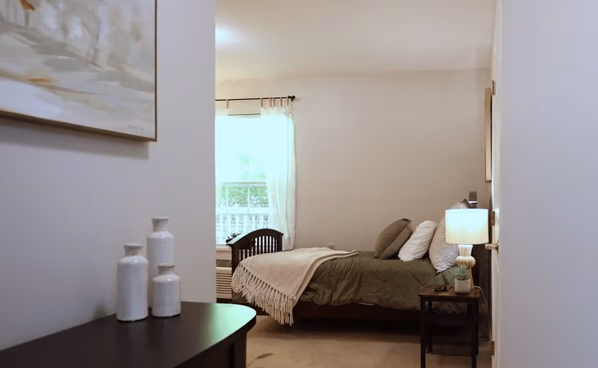 A cozy bedroom with a neatly made bed featuring green bedding and multiple pillows. A small bedside table with a lamp and a small plant is next to the bed. A window with sheer white curtains allows natural light to brighten the room. Three white decorative vases are placed on a dark wooden surface in the foreground.