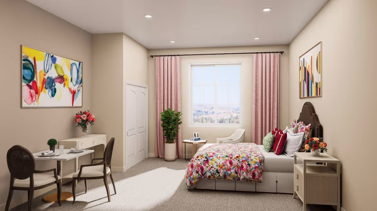 Sunlit bedroom in a senior living suite featuring a floral bed with pillows, a nightstand, seating by a window with pink curtains, a small dining table and colorful wall art.