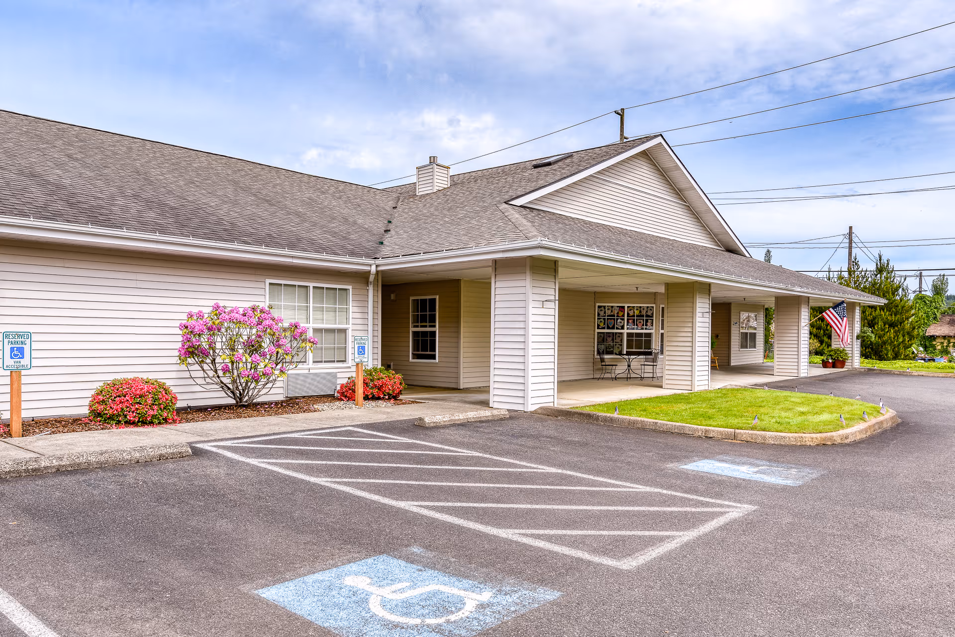 Exterior view of Riverside Place Memory Care facility showing a single-story building with beige siding, a covered entrance, a small lawn area, and a parking lot with designated handicapped parking spaces. There are flowering bushes near the building and an American flag displayed near the entrance.