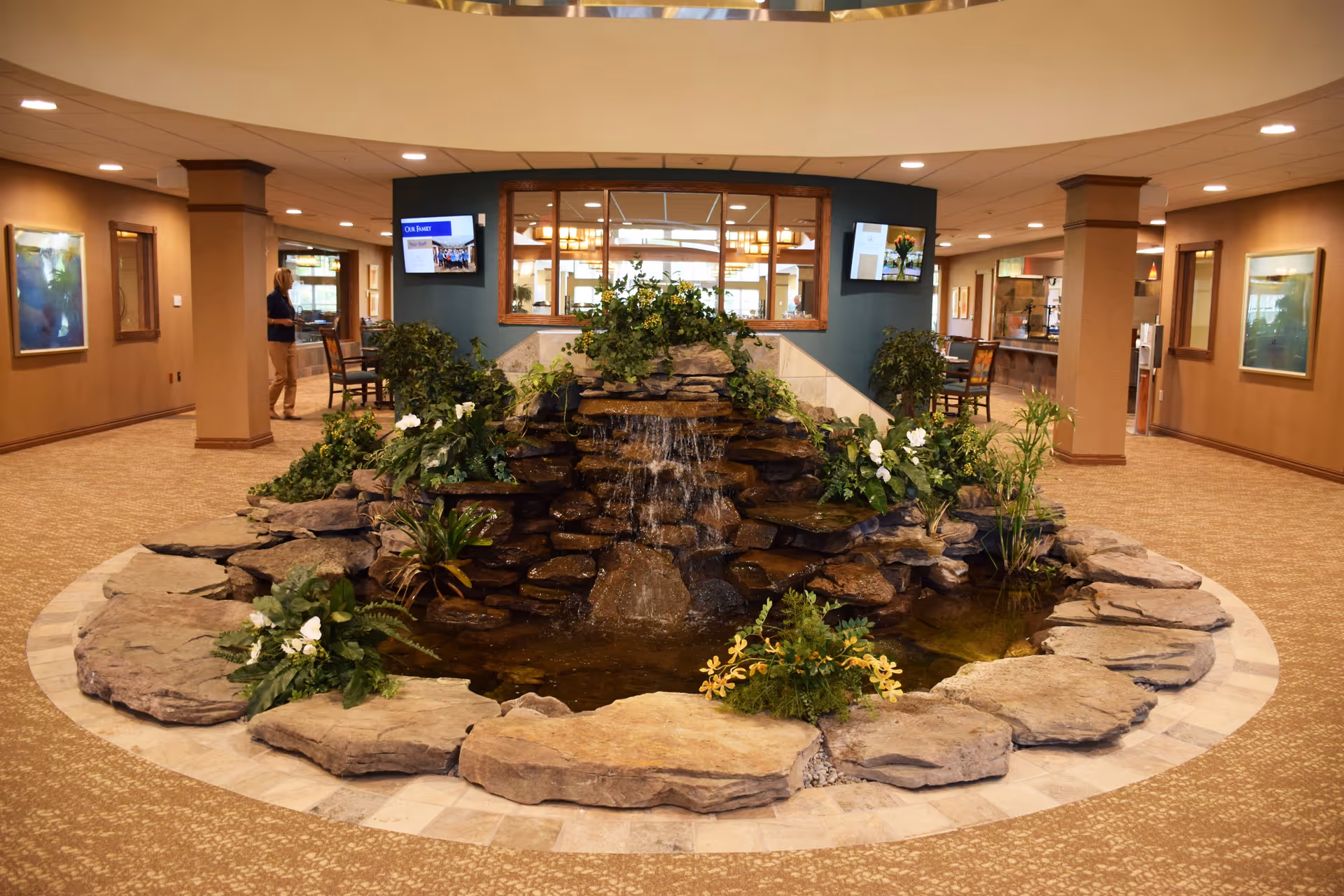 Indoor water feature with a small waterfall surrounded by rocks and plants in the center of a spacious, carpeted common area with beige walls and ceiling lights. Two flat-screen TVs are mounted on a blue accent wall behind the water feature. A woman is standing near the left side of the room, and there are chairs and tables visible in the background.