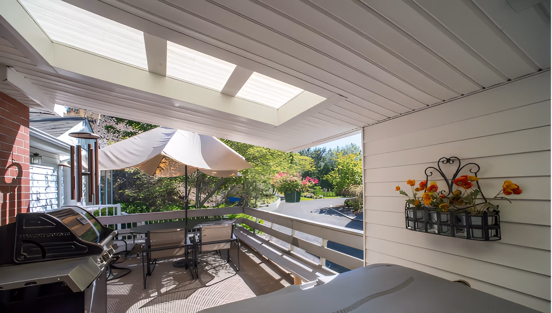 Covered outdoor patio area with a skylight, a barbecue grill, a table with chairs, a large umbrella, and a wall-mounted flower holder with orange and yellow flowers. The patio overlooks a driveway and a garden with green trees and flowering plants.