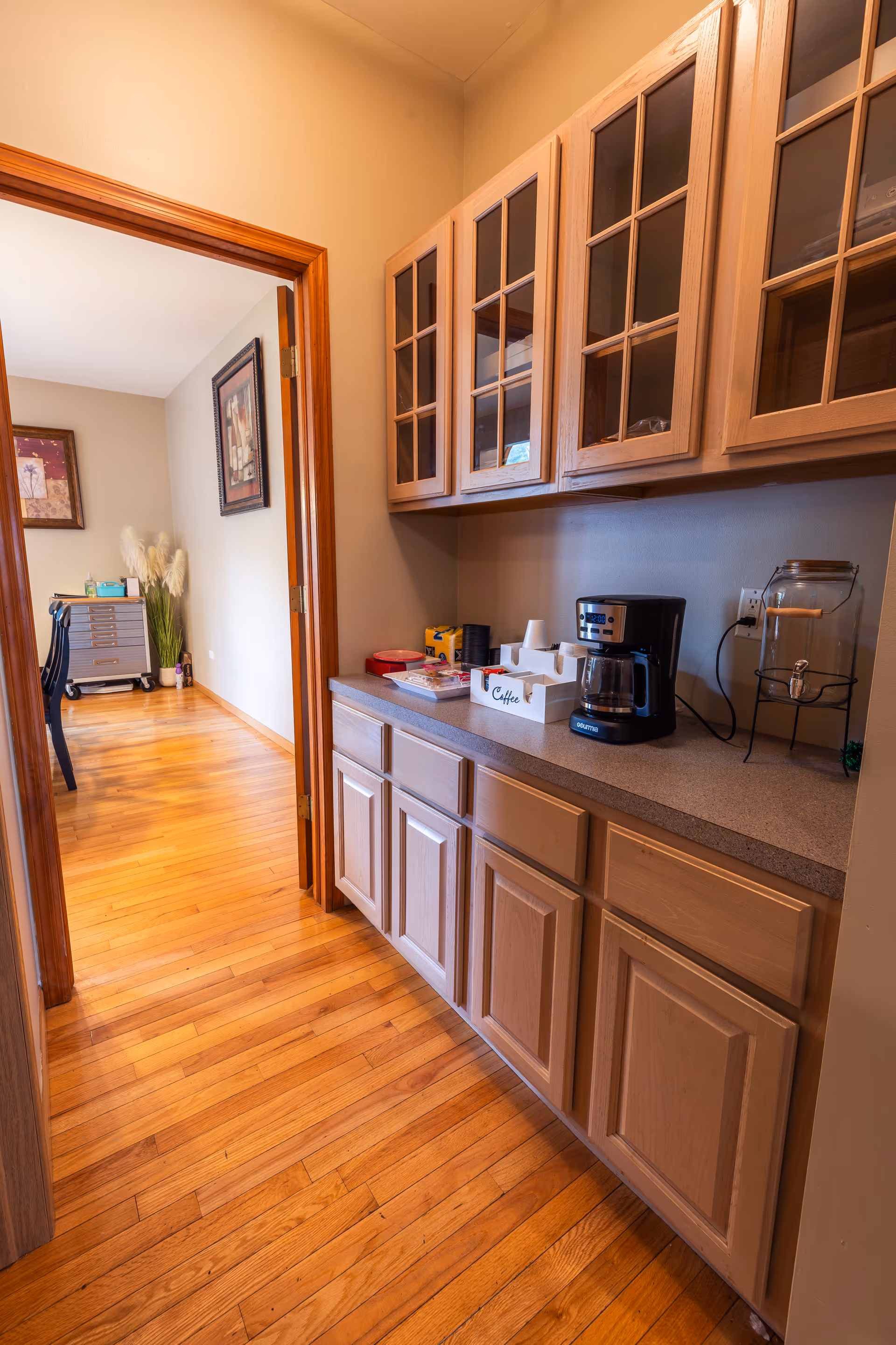 A hallway with wooden cabinets on the right side, featuring glass-front upper cabinets and solid lower cabinets. On the countertop, there is a coffee maker, a glass beverage dispenser, and a white container labeled 'Coffee' holding coffee supplies. The floor is hardwood, and an adjacent room with a desk, chair, and framed artwork is visible through an open doorway.