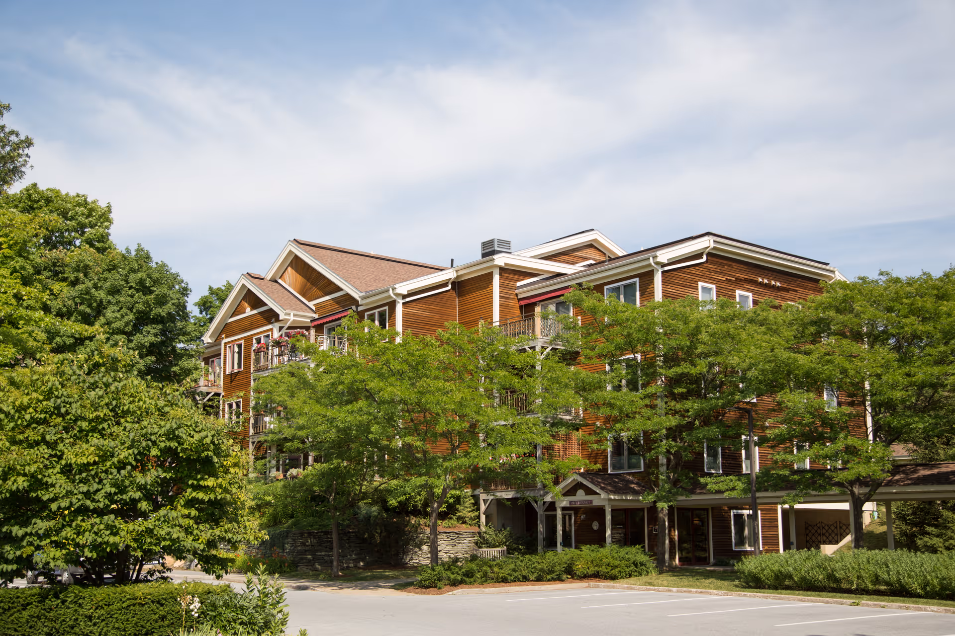 Exterior view of a multi-story wooden building surrounded by green trees under a partly cloudy sky, with a parking area in the foreground.