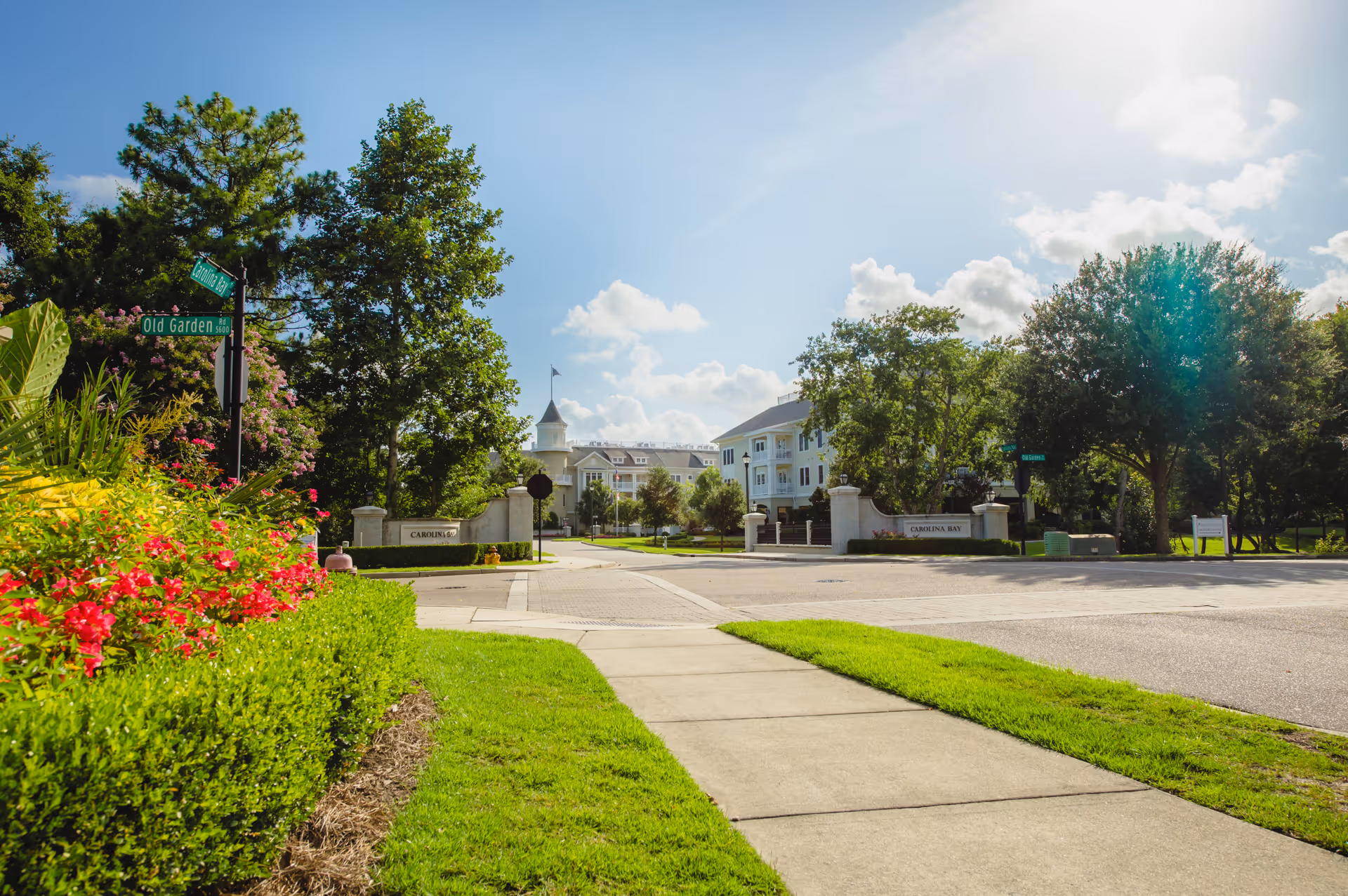View of the entrance to Carolina Bay at Autumn Hall senior living facility on a sunny day, showing a paved sidewalk, green grass, colorful flowers, trees, and the facility buildings in the background with clear blue sky and some clouds.