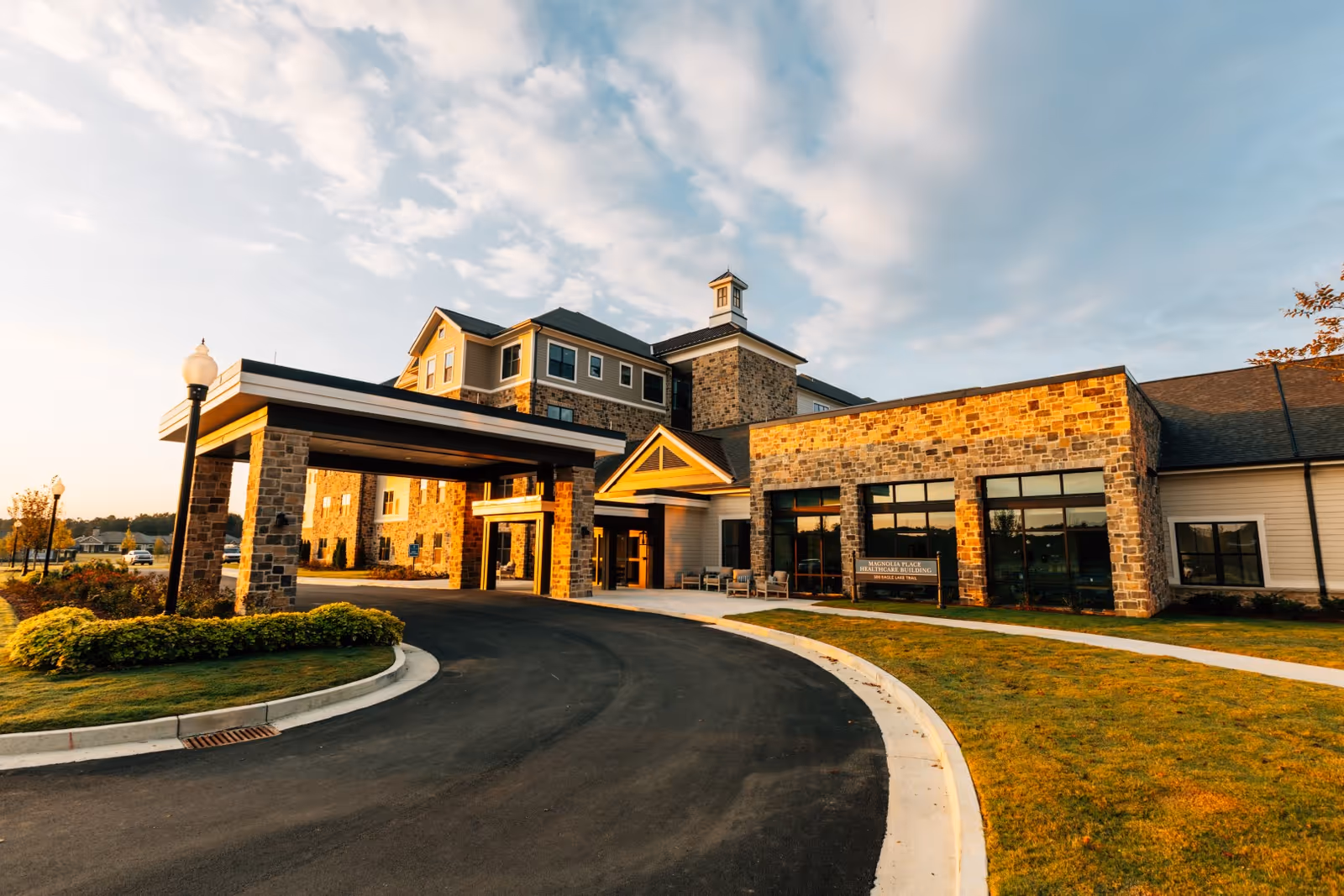 Exterior view of a senior living facility building with stone and beige siding, featuring a covered entrance with stone pillars, a curved driveway, and well-maintained landscaping under a partly cloudy sky during sunset.