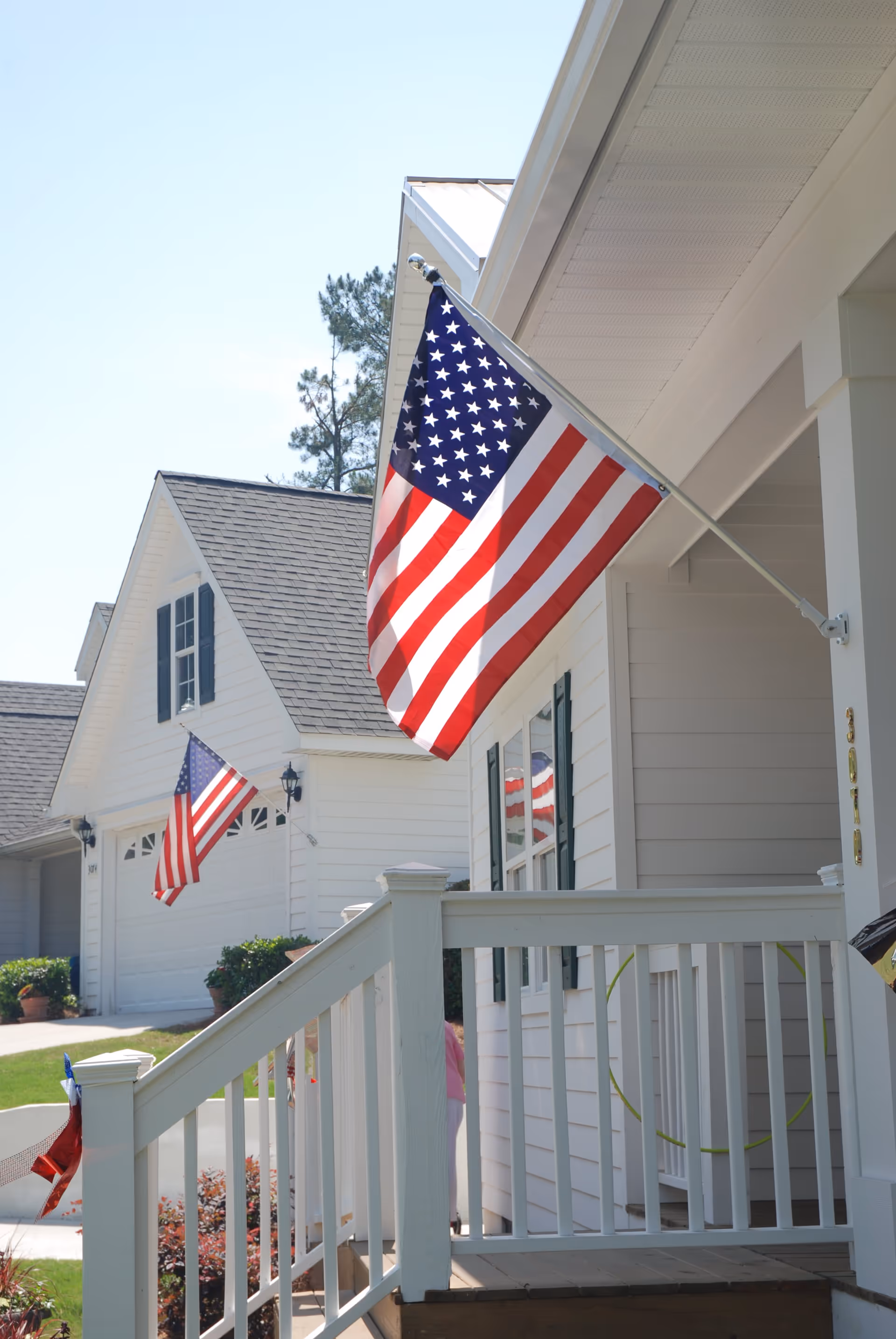 View of the front porch of a white house with an American flag mounted on a pole. Another house with a similar flag is visible in the background. The porch has white railings and steps leading up to the entrance. The sky is clear and blue.