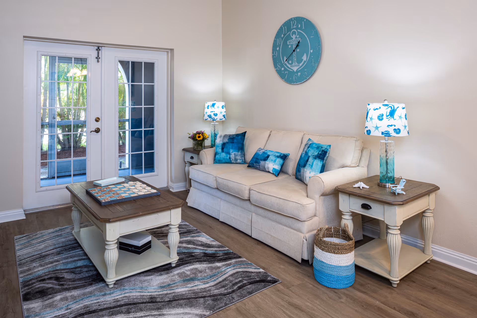 Sunlit living room with a beige sofa, blue-accent pillows, coffee table, side tables with lamps, and French doors.