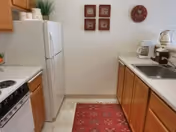 A small kitchen with wooden cabinets, a white refrigerator, a white stove, a coffee maker, a toaster, and a sink. There is a red patterned rug on the floor and four framed pictures and a clock on the wall.