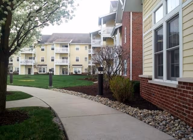 Curved concrete walkway bordered by grass, bushes, and small rocks leading through a landscaped outdoor area between multi-story residential buildings with yellow siding and brick accents under a partly cloudy sky.