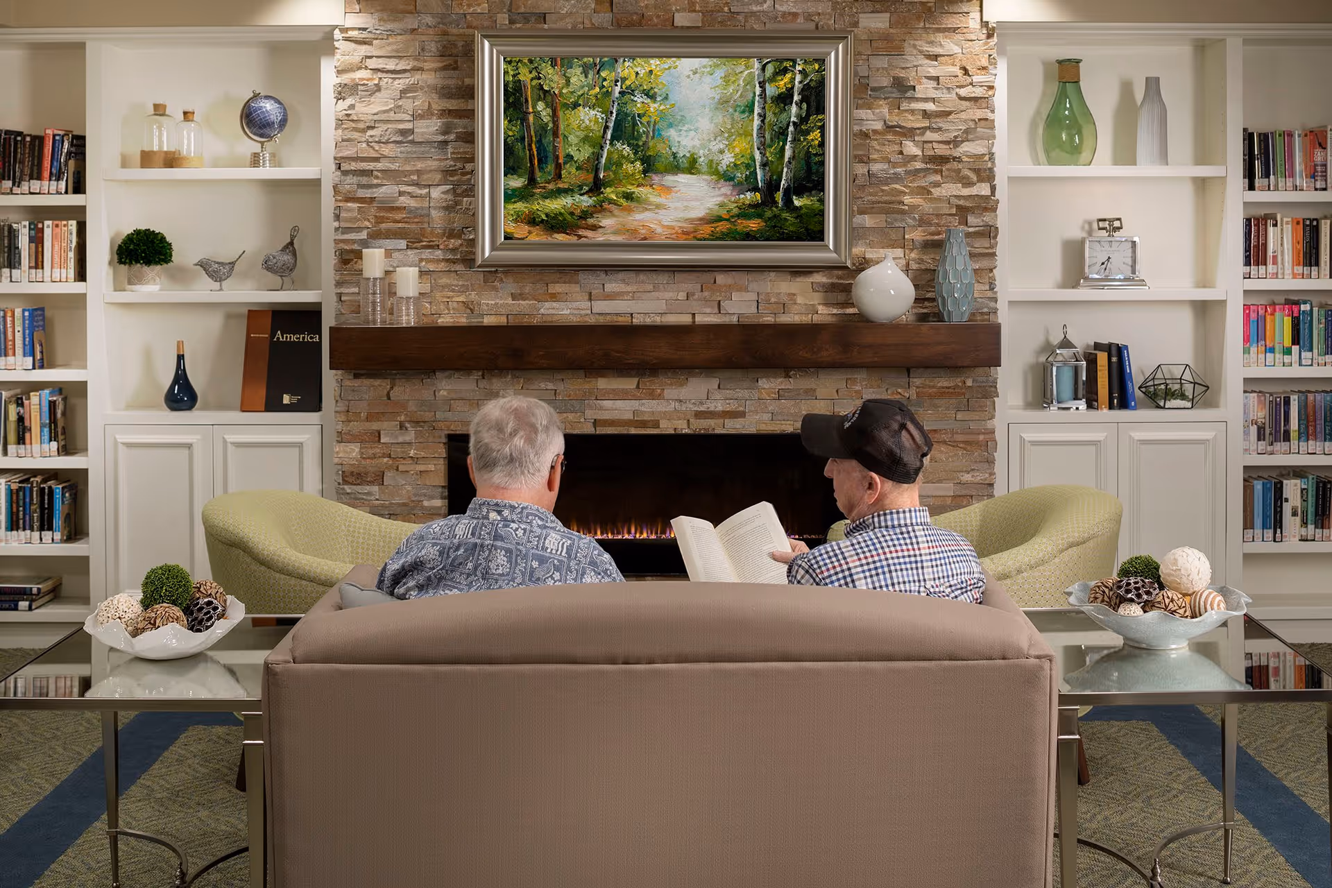 Two elderly men sitting on a beige couch facing a stone fireplace with a painting of a forest path above it. They are in a cozy room with built-in white bookshelves filled with books and decorative items. There are two green armchairs on either side of the fireplace and glass side tables with decorative bowls.