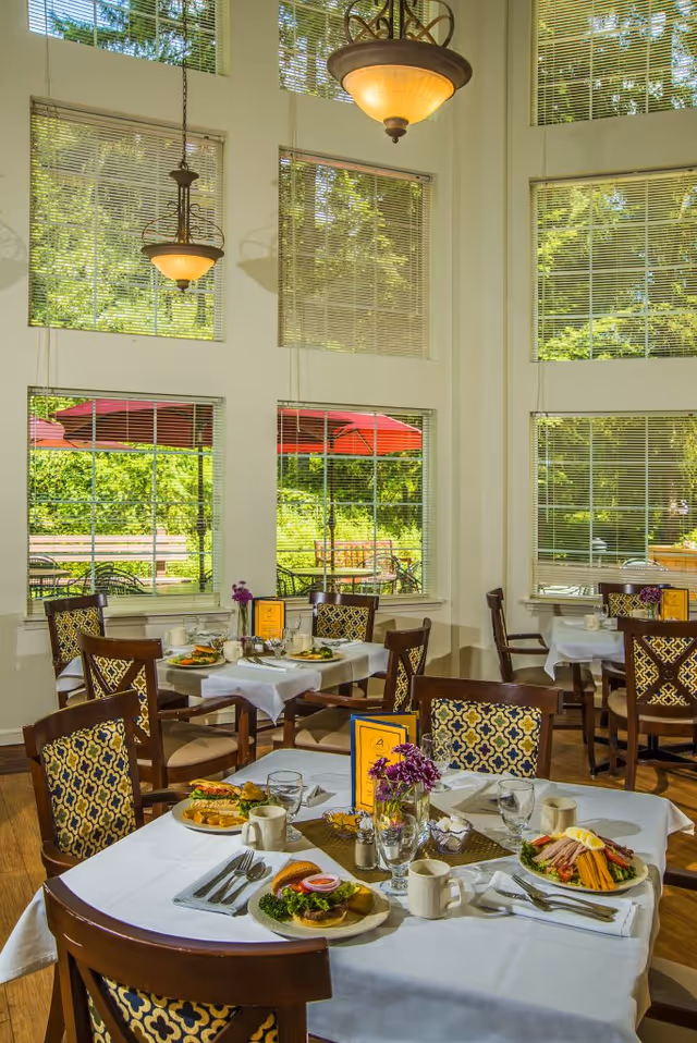 A bright dining room with large windows and white tablecloth-covered tables set with plates of food, cups, glasses, and silverware. The chairs have patterned upholstery, and there are hanging light fixtures above. Outside the windows, red umbrellas and greenery are visible.