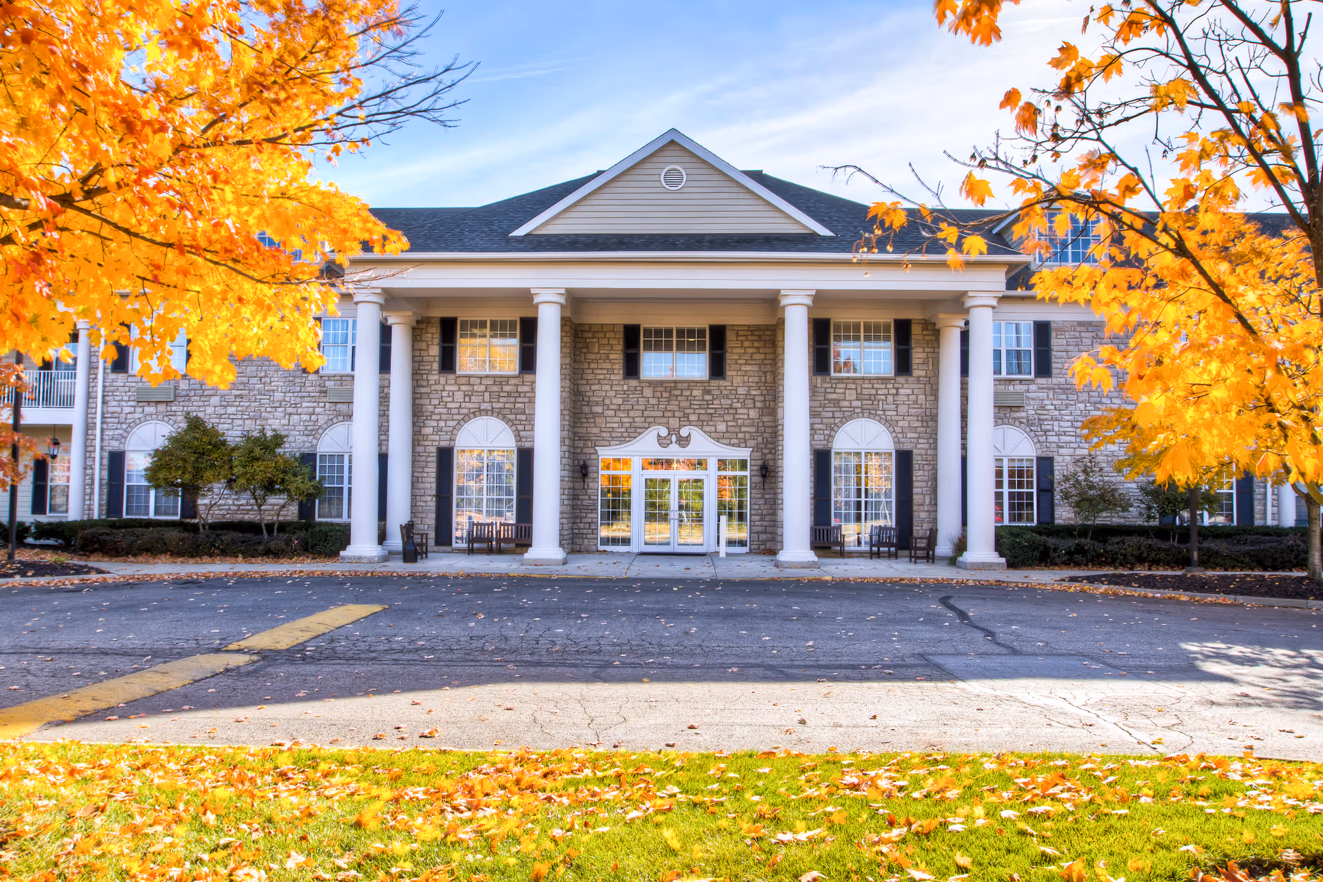 Front exterior view of a senior living facility building with stone facade, white columns, and large windows. The scene is framed by trees with bright yellow autumn leaves and fallen leaves scattered on the grass and pavement.