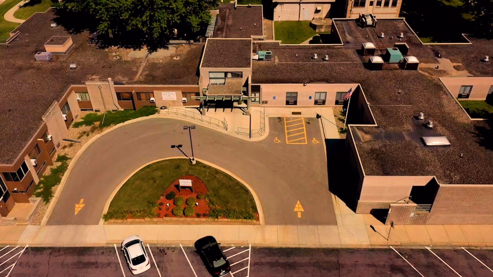 Aerial view of the front entrance of The Emeralds at Faribault facility showing a curved driveway with two handicap parking spaces, a small landscaped island with plants and a sign, and the building's flat roof and exterior walls.