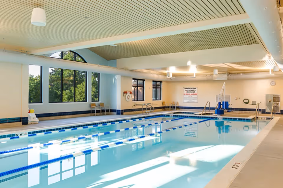 Indoor swimming pool with clear blue water, lane dividers, and a pool lift. The room has large windows letting in natural light, chairs along the walls, and a sign indicating maximum spa occupancy and no diving allowed.