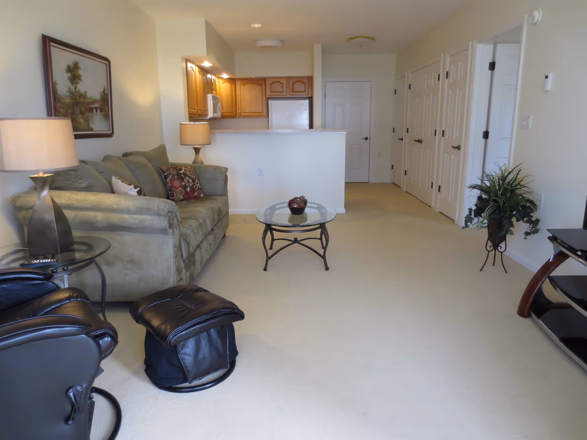 Interior view of a living room area with a green sofa, a black leather recliner and ottoman, a glass coffee table, two table lamps, a framed landscape painting on the wall, and a kitchen with wooden cabinets and white appliances in the background.