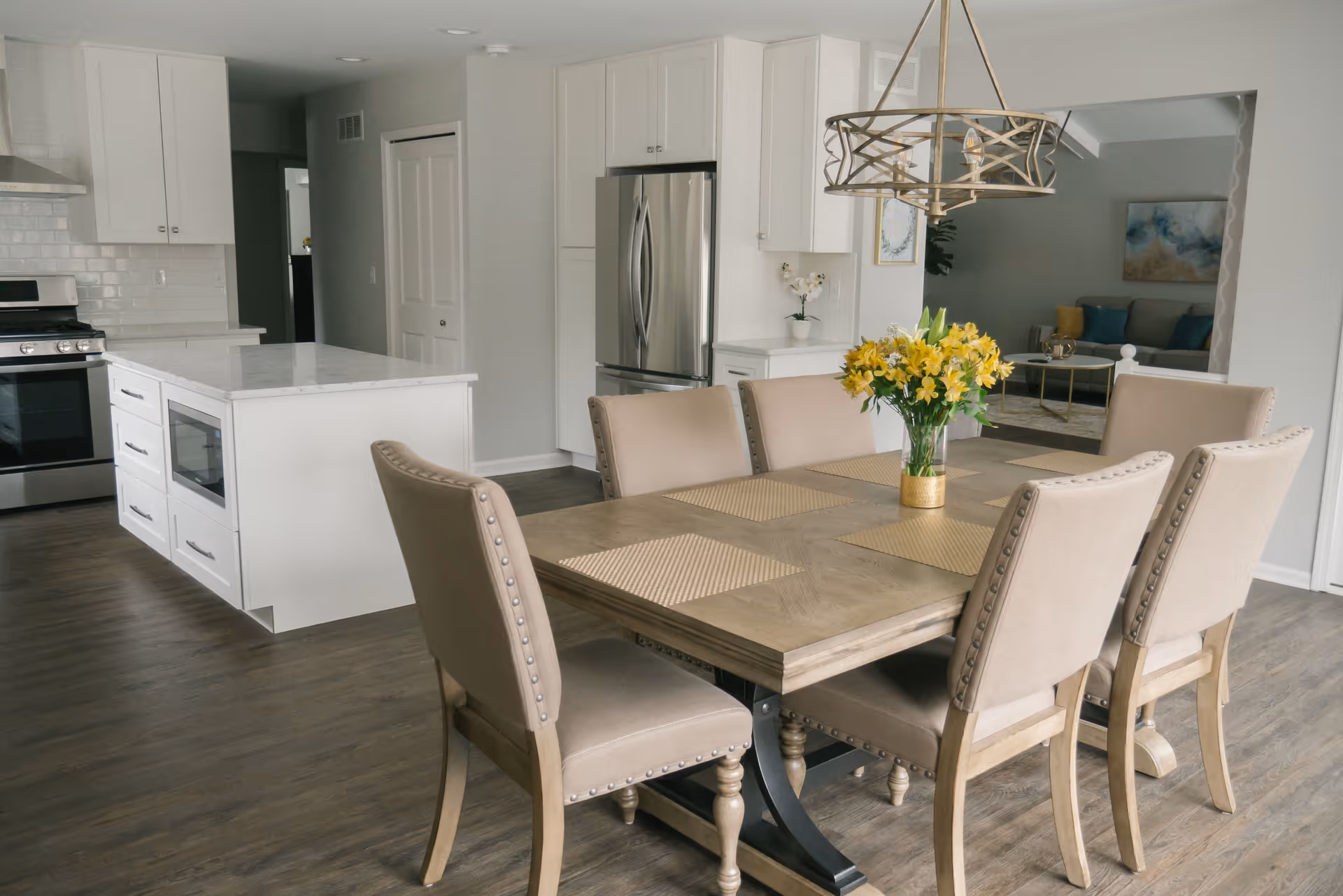 A modern dining area with a wooden table set for six with beige upholstered chairs. A vase with yellow flowers is placed in the center of the table. The dining area is adjacent to a kitchen with white cabinetry, stainless steel appliances including a refrigerator and stove, and a kitchen island with a built-in microwave. In the background, a living room with a sofa and decorative cushions is partially visible.