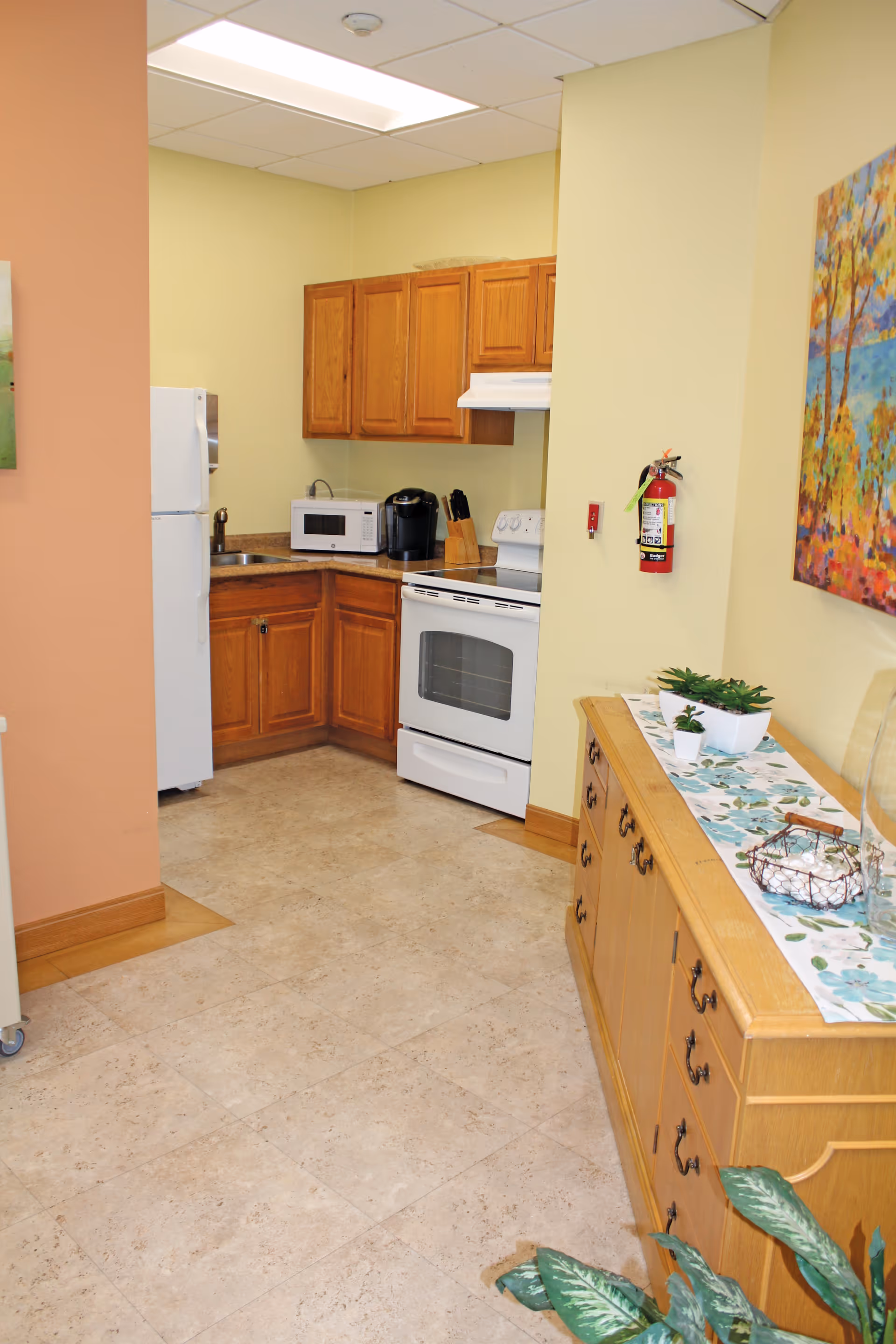 A small kitchen area with wooden cabinets, a white refrigerator, microwave, coffee maker, stove, and a sink. Adjacent to the kitchen is a wooden sideboard with decorative plants and a colorful painting on the wall above it. The floor is tiled and the walls are painted in light yellow and peach tones.