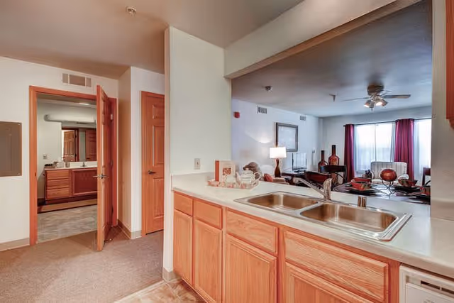 Interior view of a senior living facility apartment at The Belcaro showing a kitchen area with a double sink and wooden cabinets. Beyond the kitchen is a living room with a ceiling fan, a window with red curtains, a lamp, and a TV on a stand. To the left, an open door reveals a bathroom with a large mirror and wooden vanity.