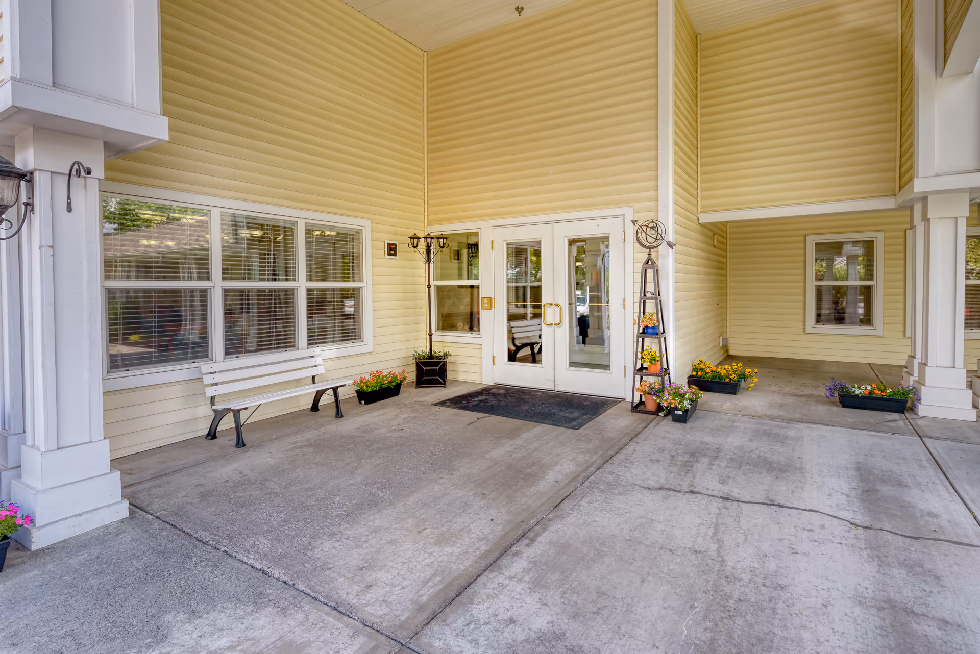 Covered entrance area of a building with yellow siding, featuring double glass doors, a white bench, several flower pots with colorful flowers, and a black doormat on a concrete floor.