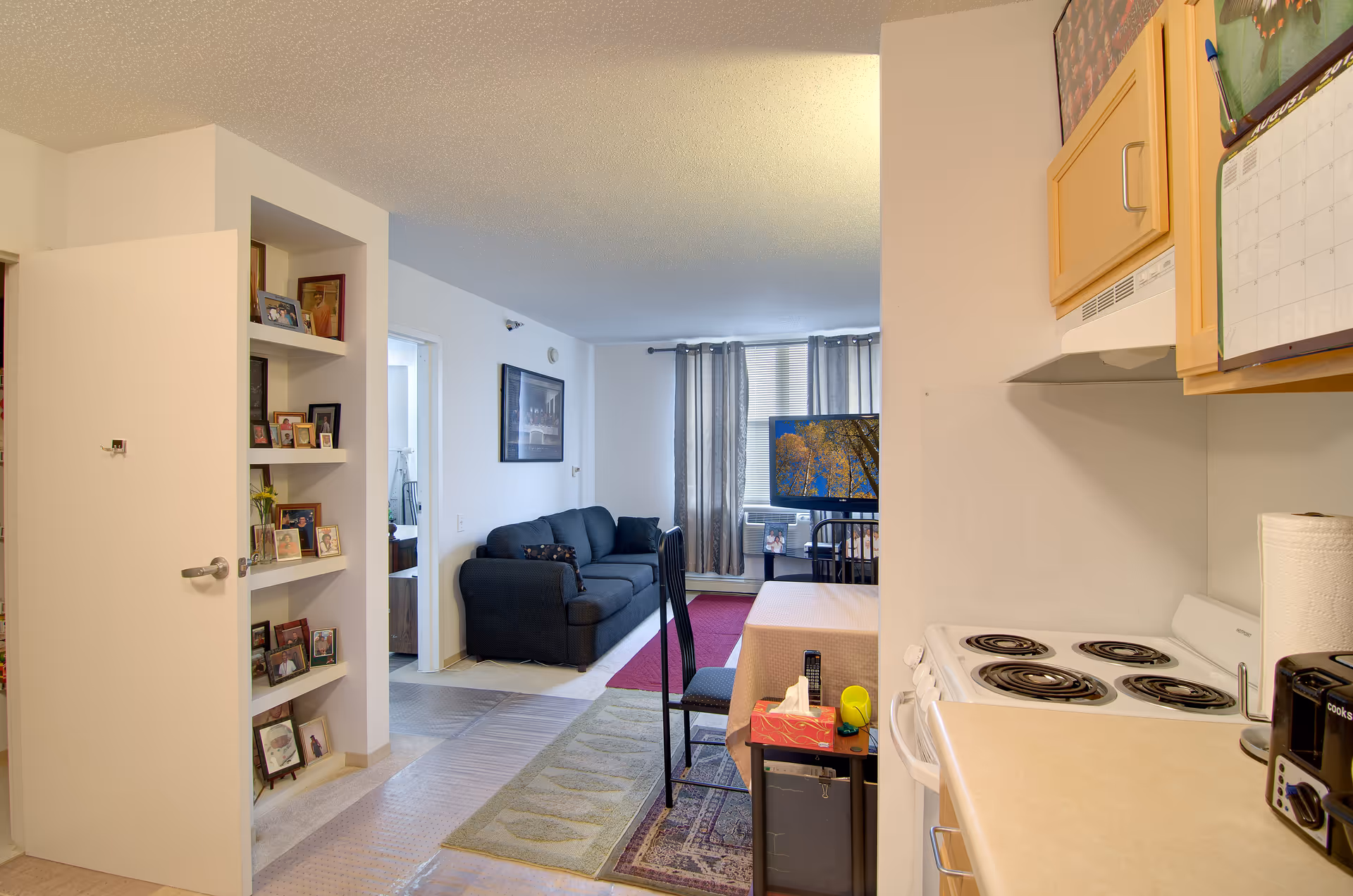 Interior view of a senior living apartment showing a small kitchen area with a stove, toaster, and paper towel holder on the right. A dining table with a chair and tissue box is adjacent to the kitchen. In the background, there is a living room with a dark blue couch, a TV on a stand, and a window with curtains. On the left, there is an open door and built-in shelves filled with framed photos.