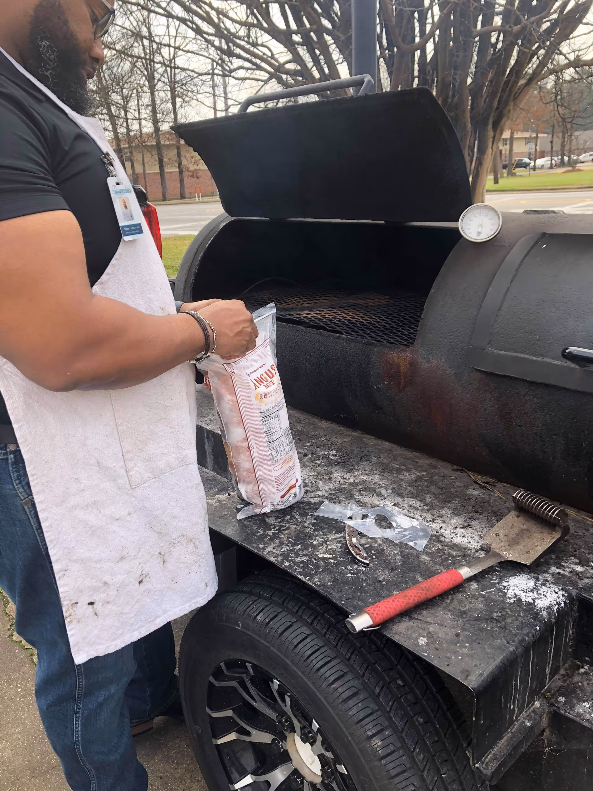 A person wearing a white apron is standing next to a large outdoor barbecue smoker grill. The person is holding a bag of wood chips or charcoal and appears to be preparing the grill. The grill lid is open, showing the cooking grate inside. There is a spatula with a red handle resting on the grill's side shelf. Trees and a street with parked cars are visible in the background.