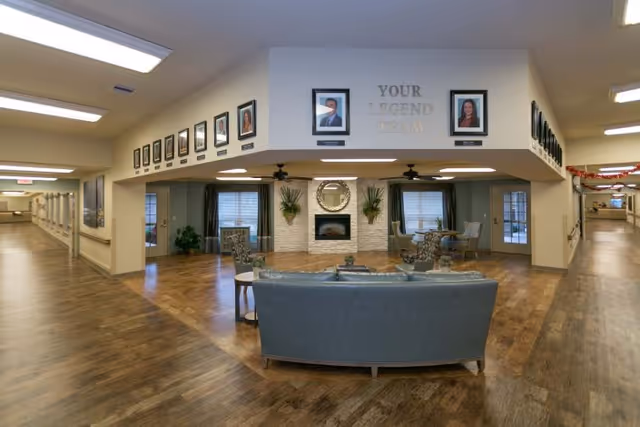 A spacious interior common area in a senior living facility with a seating arrangement including a blue sofa and chairs around a fireplace. The walls above the seating area display framed portraits with the text 'YOUR LEGEND TEAM'. The area has wood flooring and is well-lit with ceiling lights. Hallways extend on both sides of the common area.