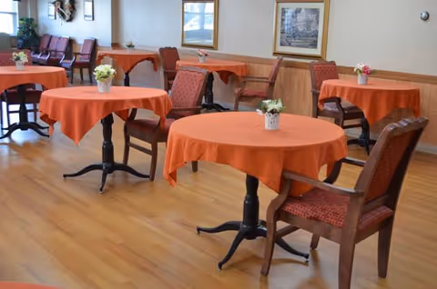 A dining area with round tables covered in orange tablecloths, each table decorated with a small flower arrangement. Wooden chairs with red patterned cushions surround the tables. The room has light-colored wooden flooring, beige walls with wood paneling, and framed pictures hanging on the walls.