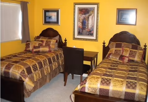 A bedroom with two twin beds featuring matching brown and yellow patterned bedding. Between the beds is a small dark wooden desk with a dark chair. The walls are painted yellow and decorated with three framed pictures. A window with closed blinds and a brown curtain is on the left side of the room.