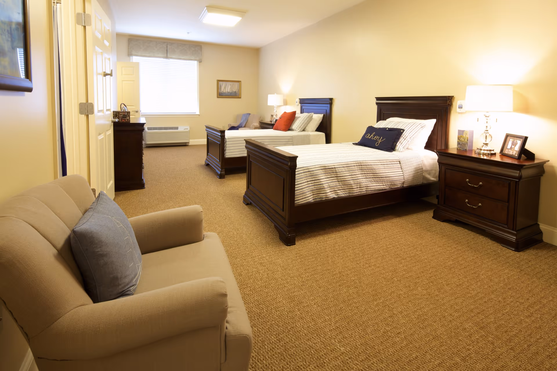 A senior living facility bedroom with two wooden twin beds, each with striped bedding and decorative pillows. There is a wooden nightstand with a lamp, picture frame, and small decorations between the beds. A beige armchair with a blue pillow is in the foreground, and a window with blinds is at the far end of the room. The walls are painted light yellow and the floor is carpeted.