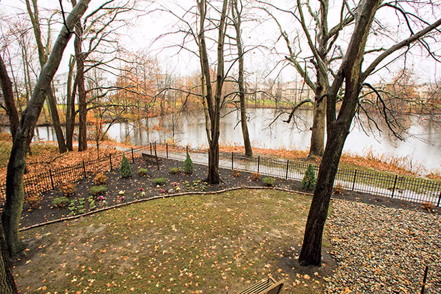 View of a fenced garden area with small plants and trees, fallen autumn leaves on the ground, a bench, and a river in the background with leafless trees along the riverbank.