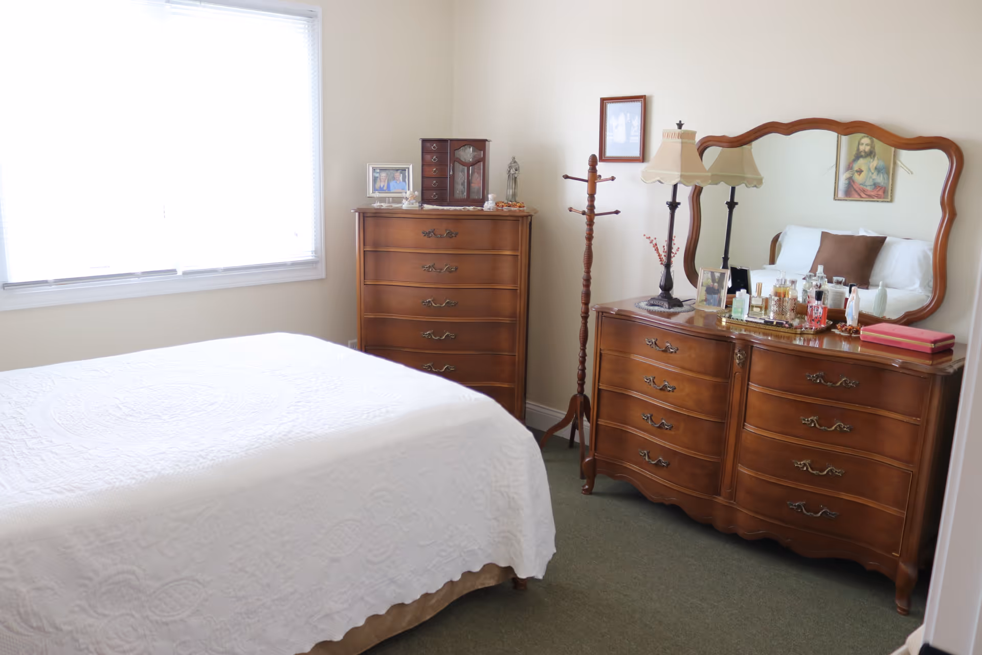 A bright bedroom with a white-covered bed, wooden dressers and mirror, a lamp, and a large window.
