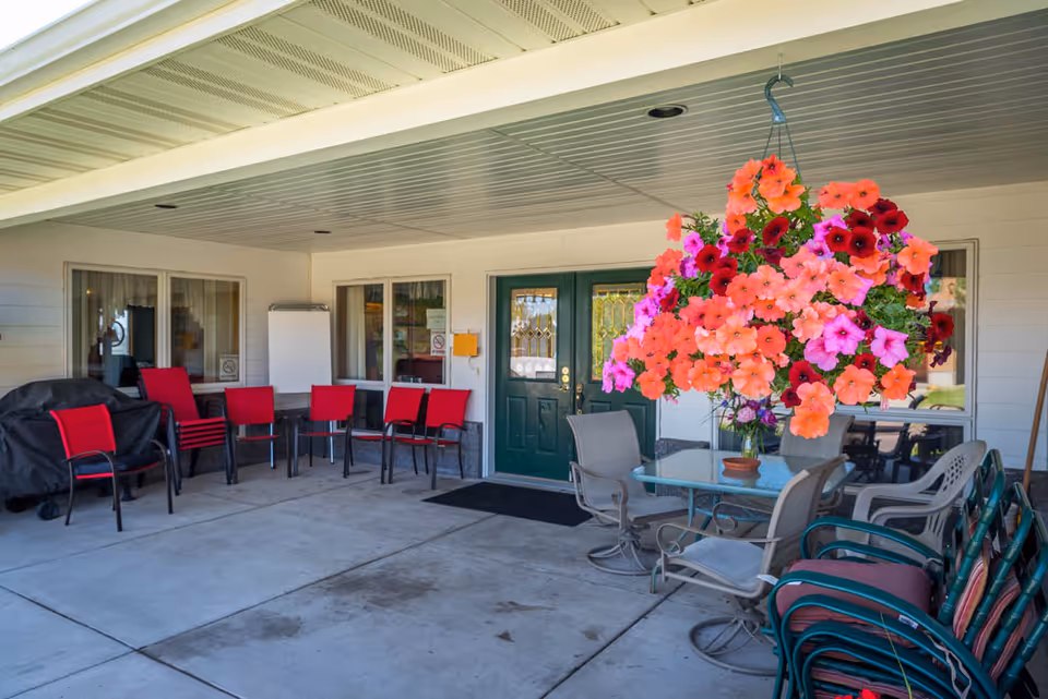 Covered entrance patio with a large hanging basket of pink and orange flowers, tables and chairs arranged near green double doors.