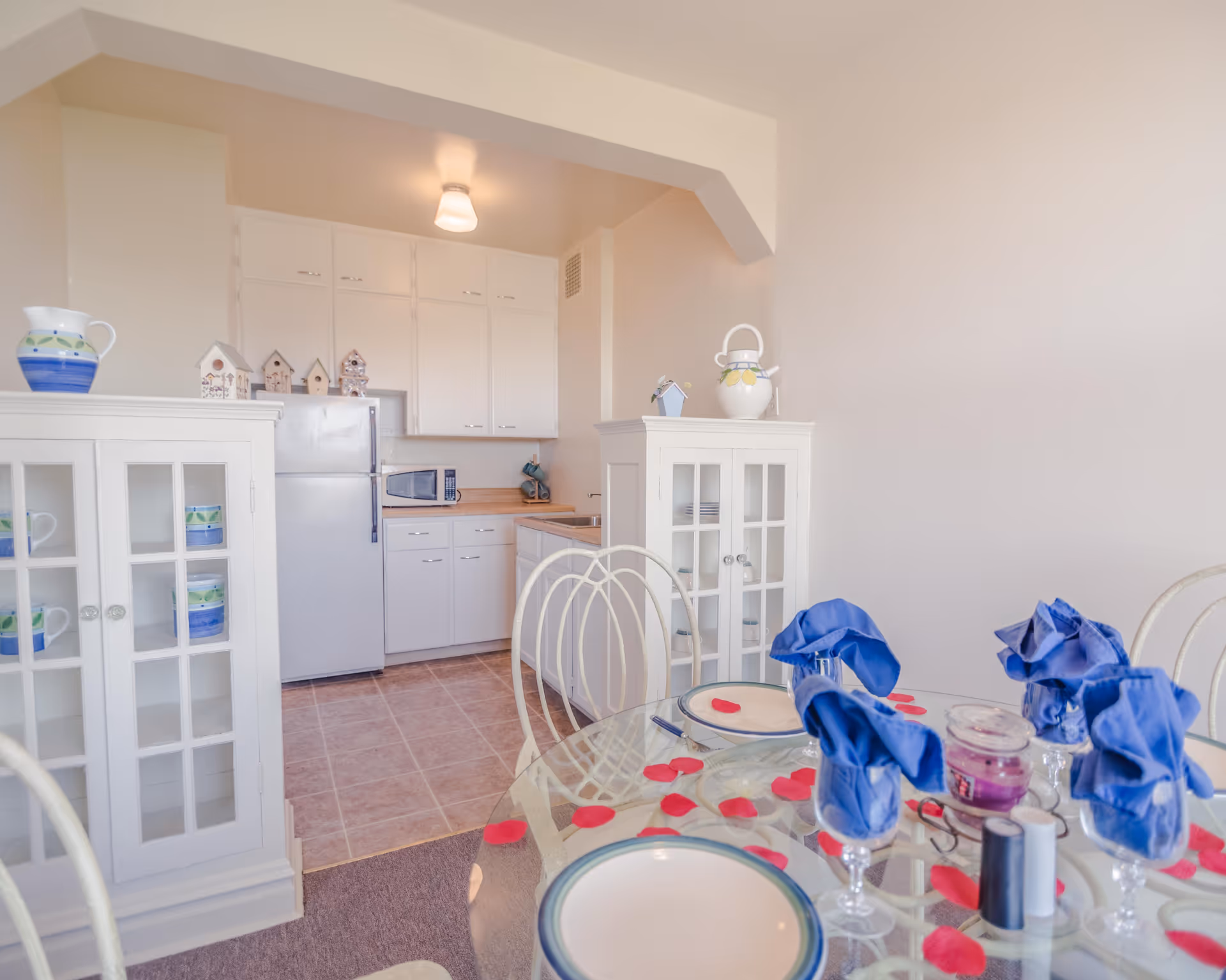 A bright kitchen and dining area with white cabinets, a refrigerator, microwave, and a glass dining table set with plates, glasses with blue napkins, and scattered red rose petals. Two white cabinets with glass doors display cups and decorative items.