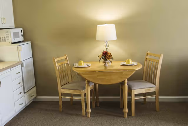 A small wooden dining table with two chairs set against a beige wall. The table has a lamp with a white shade and a small floral arrangement in the center. Two place settings with yellow cups are arranged on the table. To the left, there is a white kitchen counter with a microwave and refrigerator.