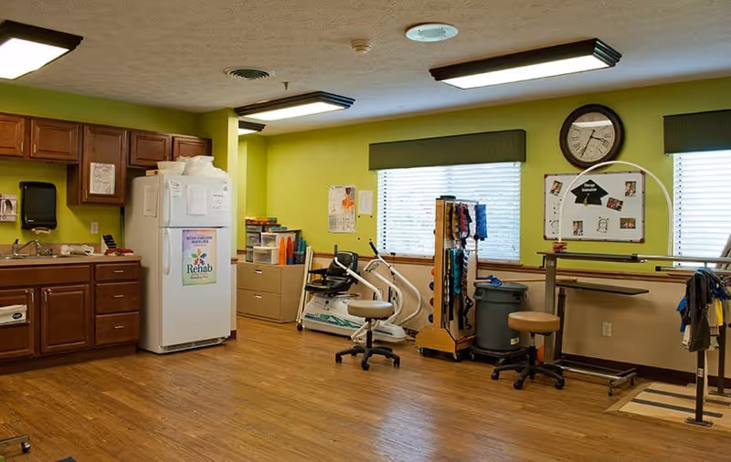 Interior rehabilitation room with a kitchenette, wooden floors, exercise equipment including a stationary bike and parallel bars, and storage cabinets.